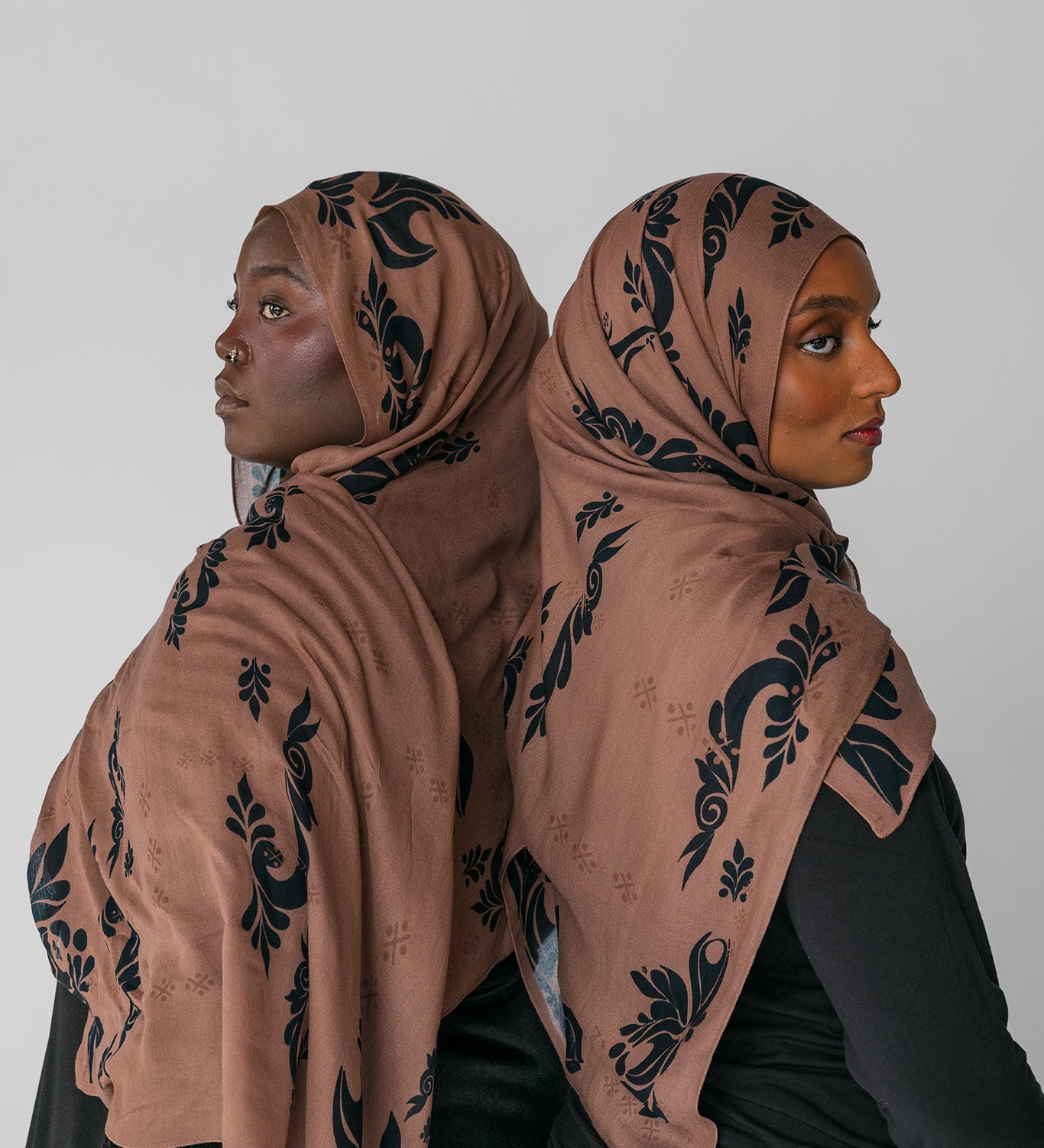 Two women wearing matching brown hijabs with black patterns against a white background