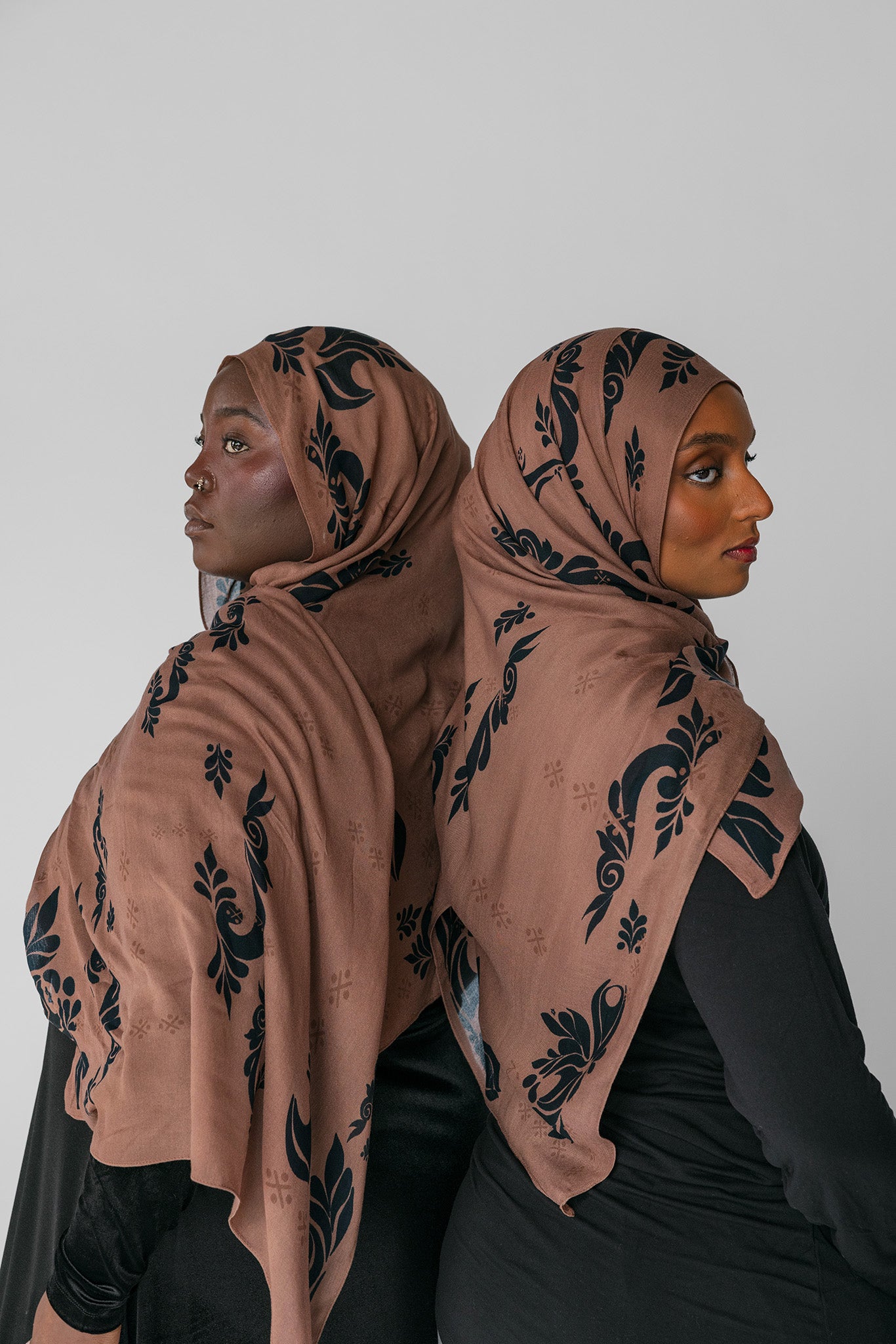 Two women wearing matching brown hijabs with black patterns against a white background
