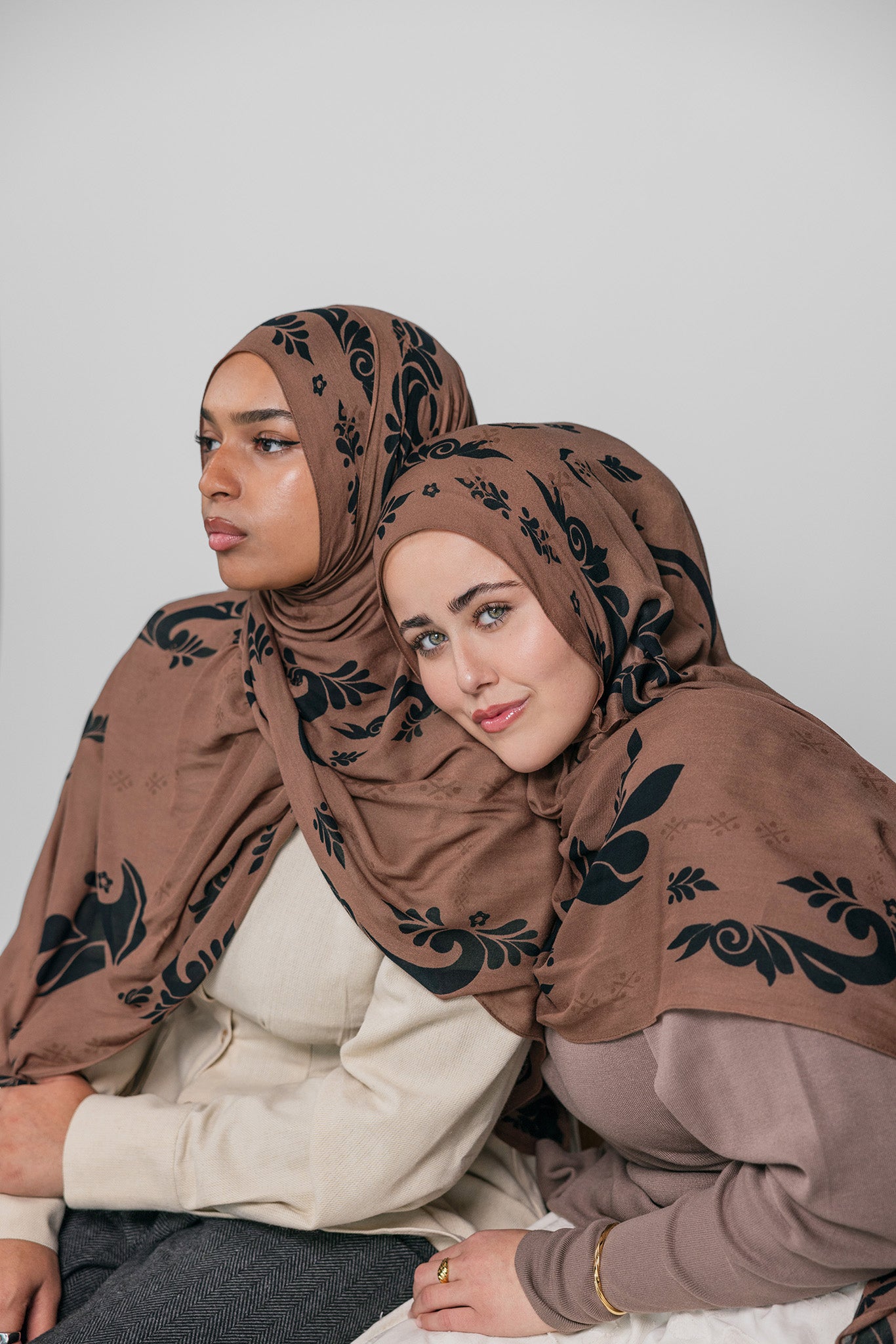 Two women wearing brown hijabs with black patterns on a white background