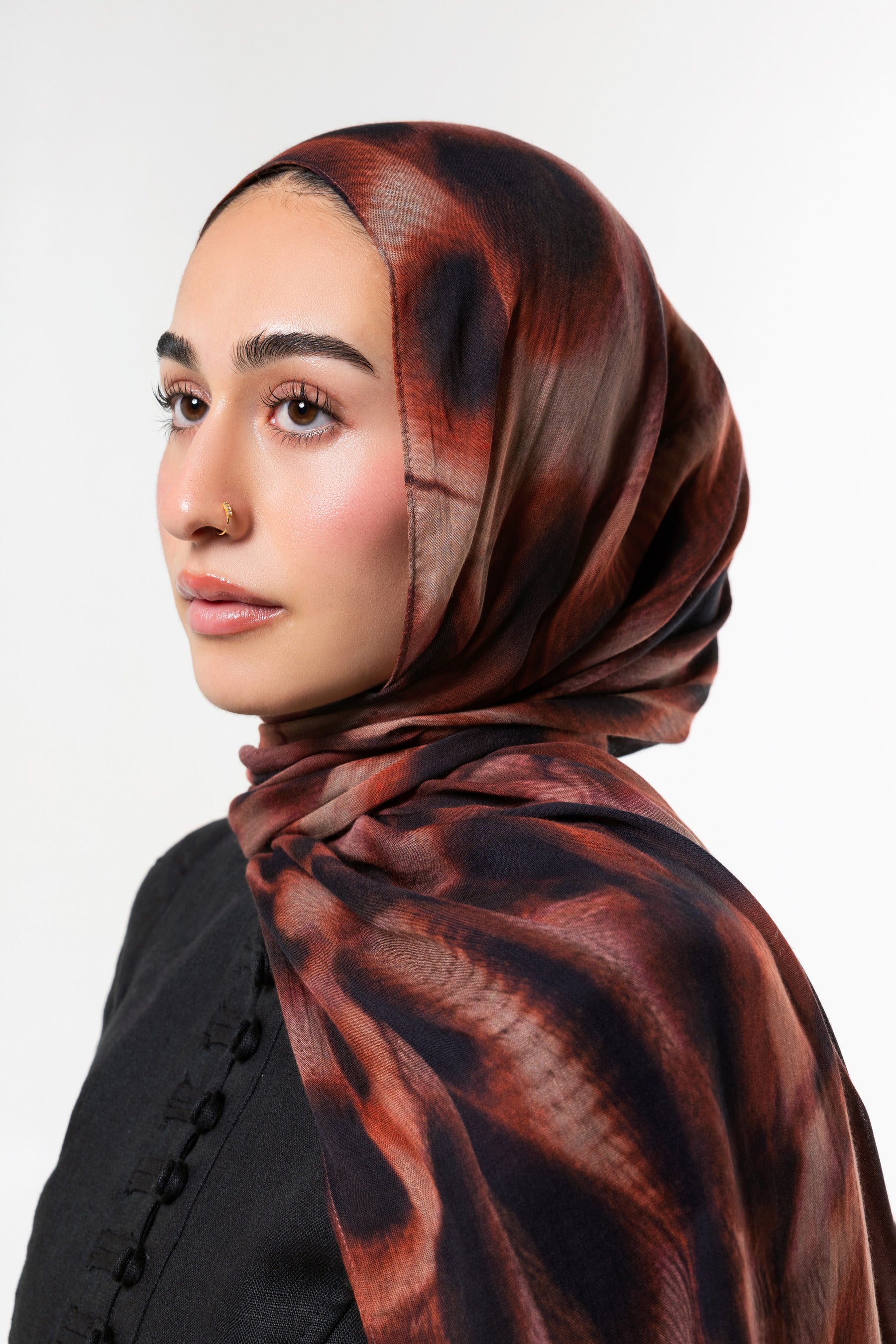 A woman wearing a dark brown and black patterned hijab, photographed in profile against a clean light gray studio background.
