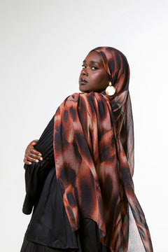 A woman wearing a black outfit and a brown and black patterned hijab, photographed against a plain light gray studio background.