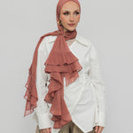 A woman wearing a nectar ruffled hijab, a white shirt, and beige trousers, standing against a light gray studio background.
