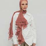 A woman wearing a nectar ruffled hijab, a white shirt, and beige trousers, standing against a light gray studio background.