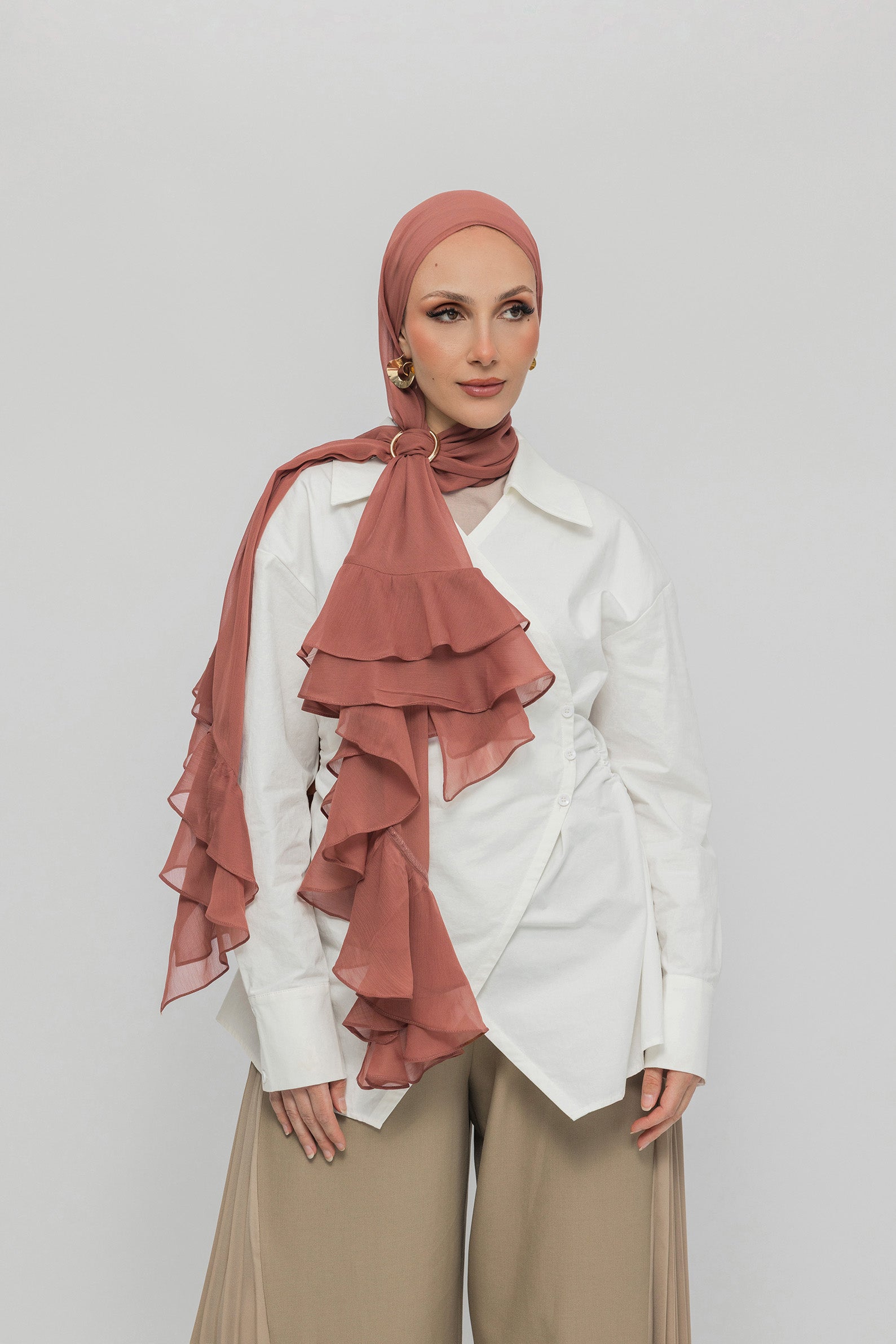A woman wearing a nectar ruffled hijab, a white shirt, and beige trousers, standing against a light gray studio background.