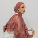 A woman wearing a nectar ruffled hijab and a white blouse with beige trousers, posed against a solid light gray background.