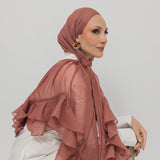 A woman wearing a nectar ruffled hijab and a white blouse with beige trousers, posed against a solid light gray background.