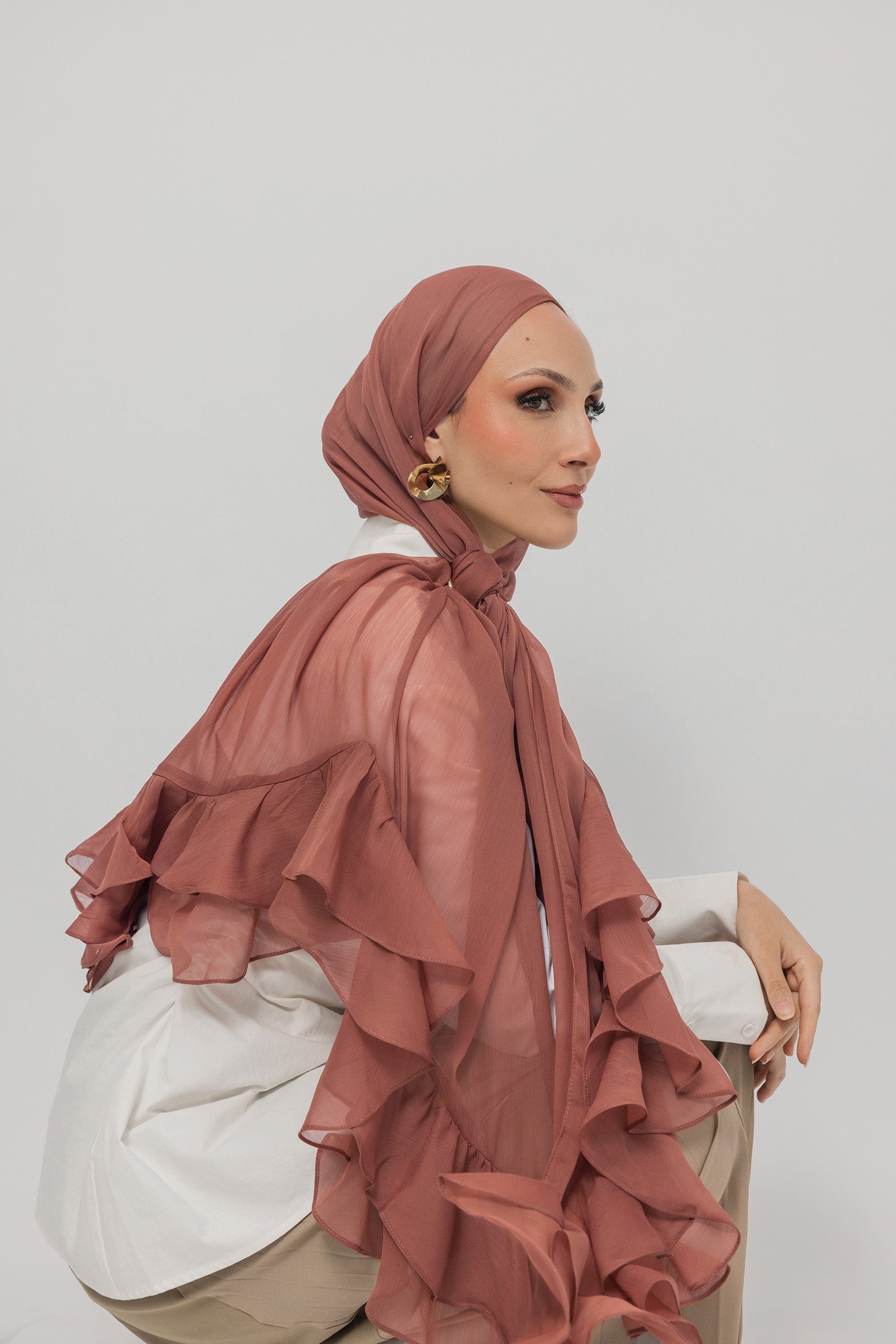 A woman wearing a nectar ruffled hijab and a white blouse with beige trousers, posed against a solid light gray background.