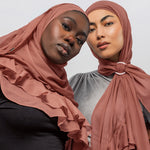 Two women wearing nectar ruffled hijabs, one draped over her shoulders and the other tied at the neck with a ring, against a light gray studio background.