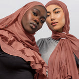 Two women wearing nectar ruffled hijabs, one draped over her shoulders and the other tied at the neck with a ring, against a light gray studio background.