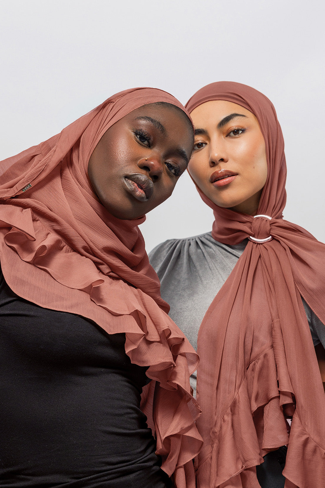 Two women wearing nectar ruffled hijabs, one draped over her shoulders and the other tied at the neck with a ring, against a light gray studio background.