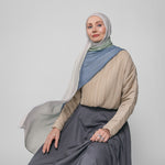Woman wearing a beige top, blue ombre hijab, and gray skirt against a plain background