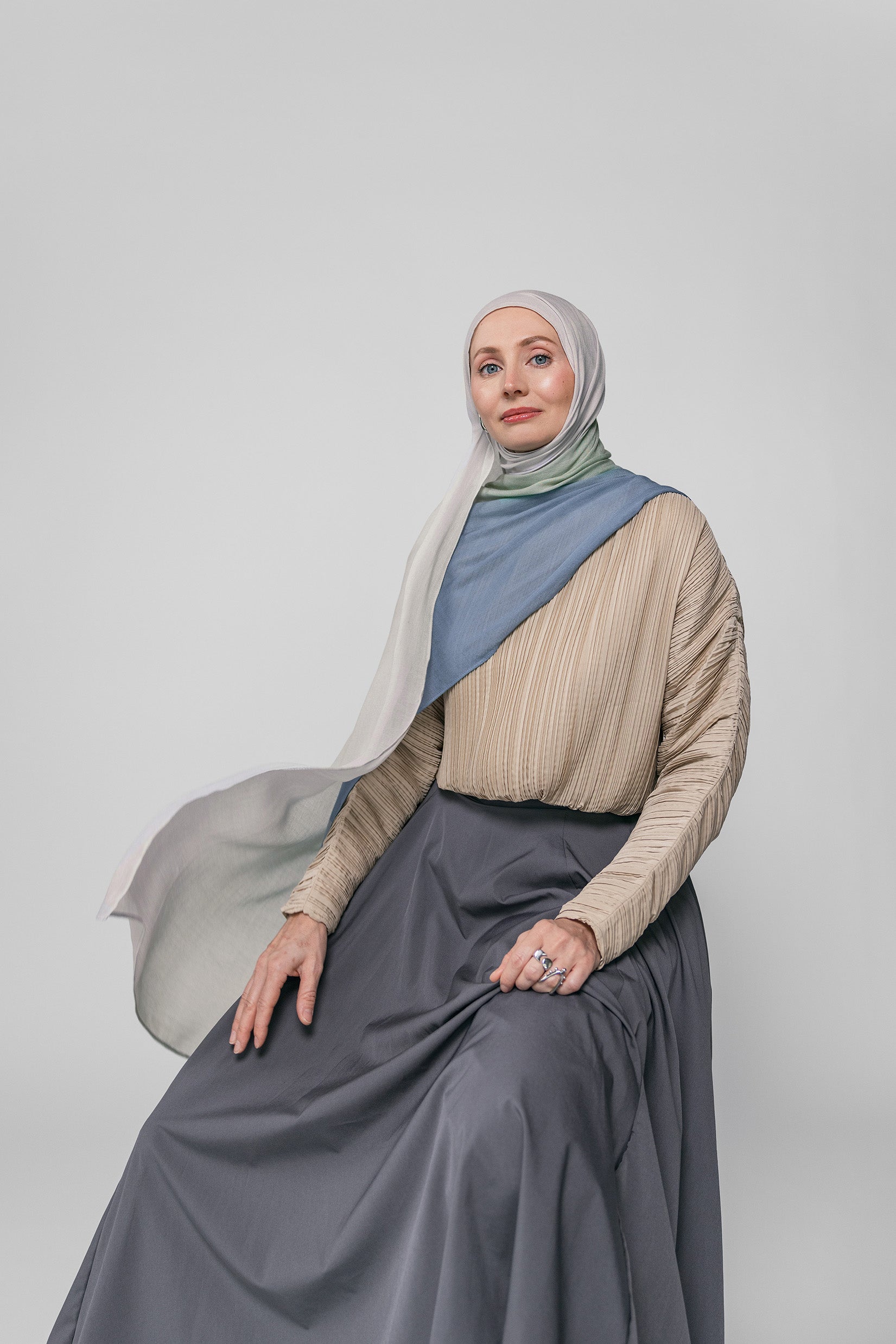 Woman wearing a beige top, blue ombre hijab, and gray skirt against a plain background