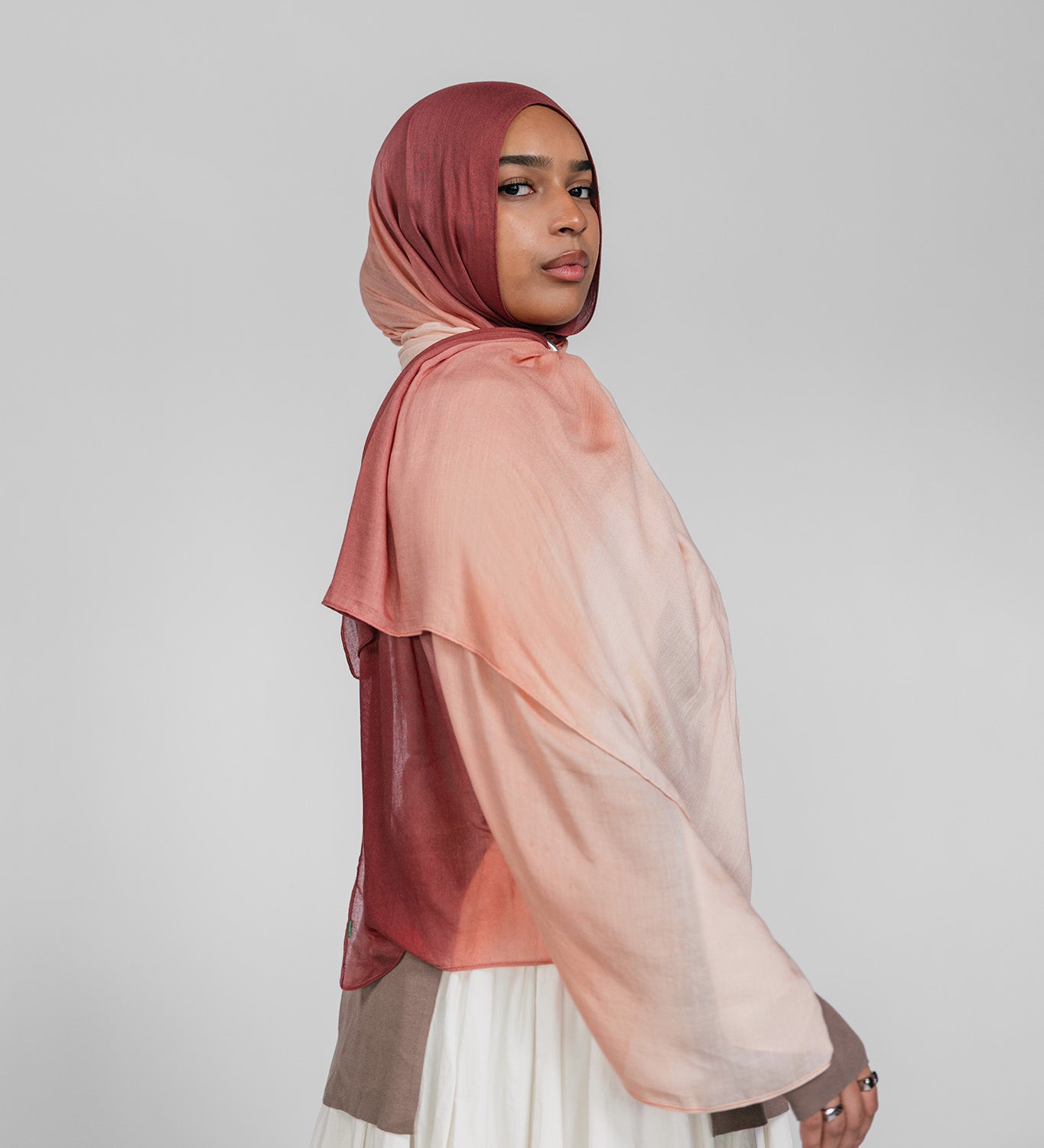 Woman wearing a brown and pink ombre hijab and white skirt on a plain background