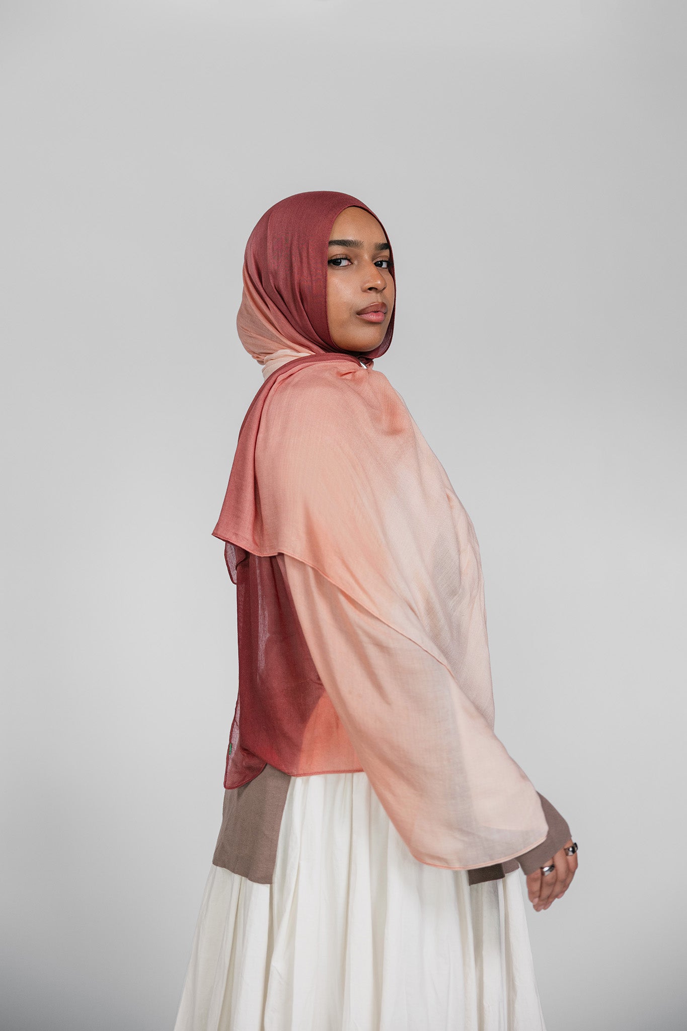 Woman wearing a brown and pink ombre hijab and white skirt on a plain background