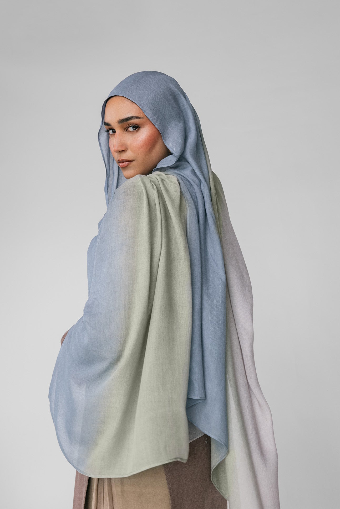 Woman wearing a light blue ombre hijab and beige outfit on a plain background