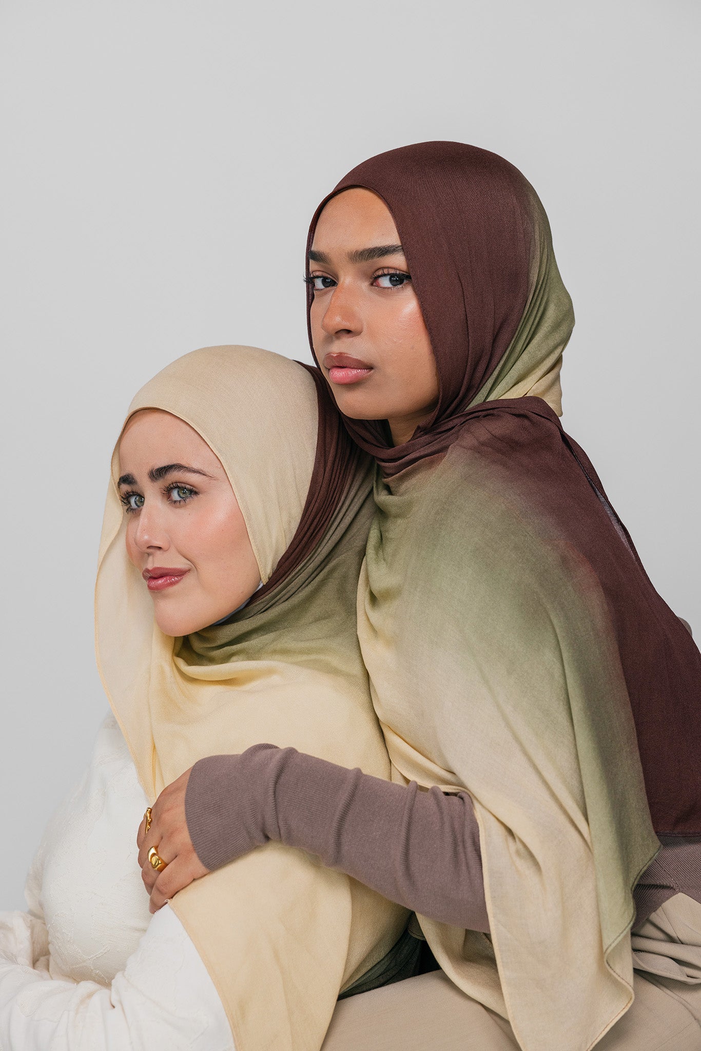 Two women wearing hijabs in beige and brown on a plain background