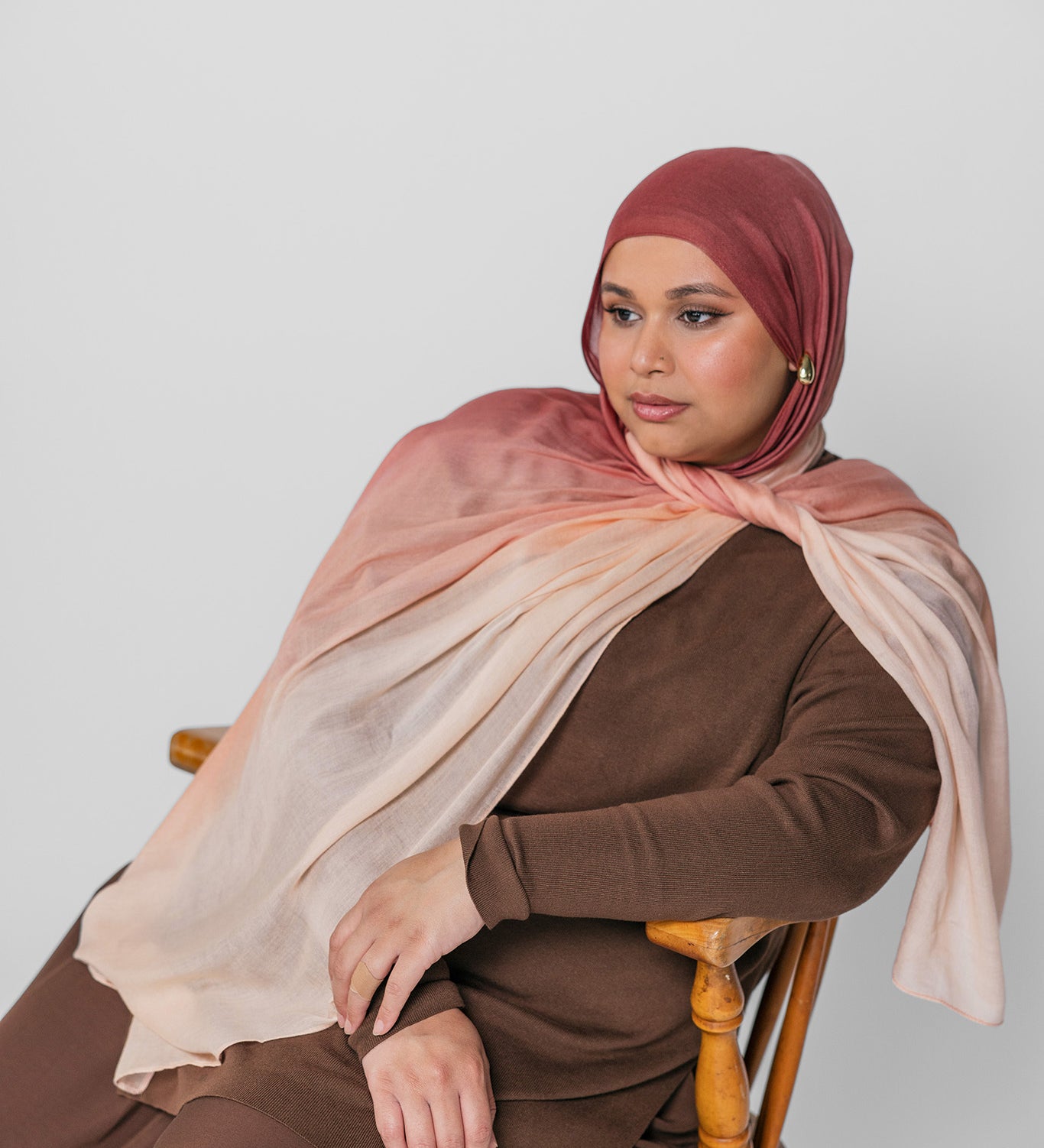 Woman wearing a brown and pink ombre hijab sitting on a wooden chair against a white background
