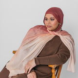 Woman wearing a brown and pink ombre hijab sitting on a wooden chair against a white background