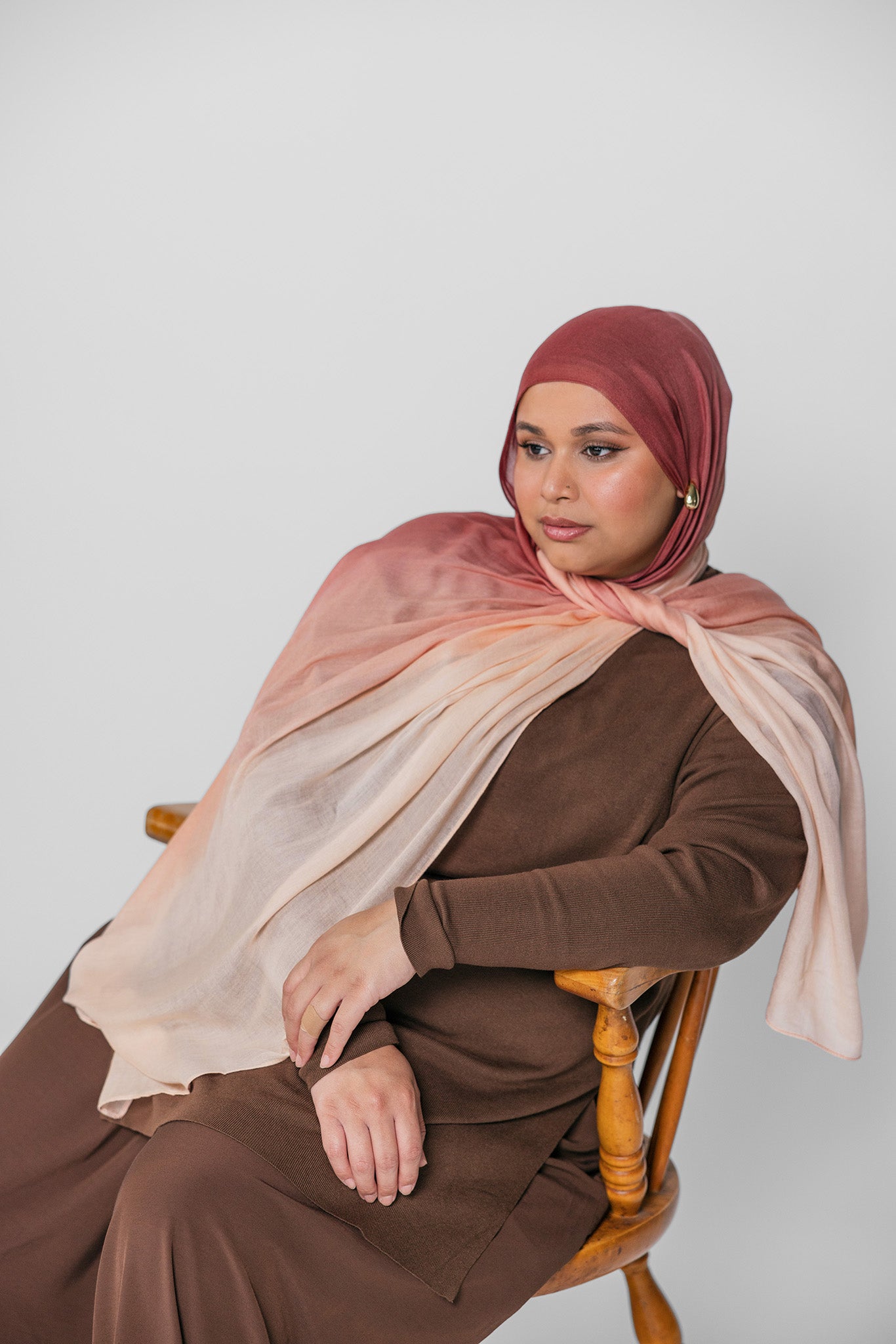 Woman wearing a brown and pink ombre hijab sitting on a wooden chair against a white background