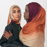 Two women wearing colorful ombre hijabs standing close together against a white background