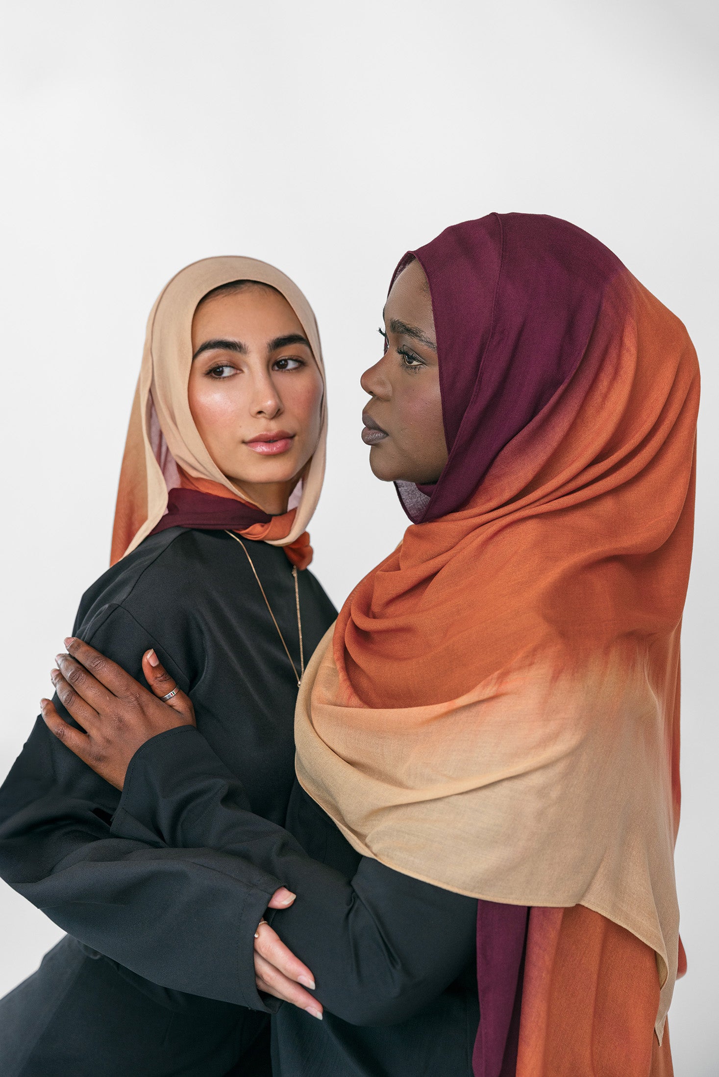 Two women wearing colorful ombre hijabs standing close together against a white background