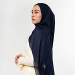 A woman in profile wearing a dark navy blue hijab and a cream-colored dress, photographed against a clean white studio background.