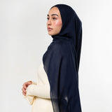 A woman in profile wearing a dark navy blue hijab and a cream-colored dress, photographed against a clean white studio background.