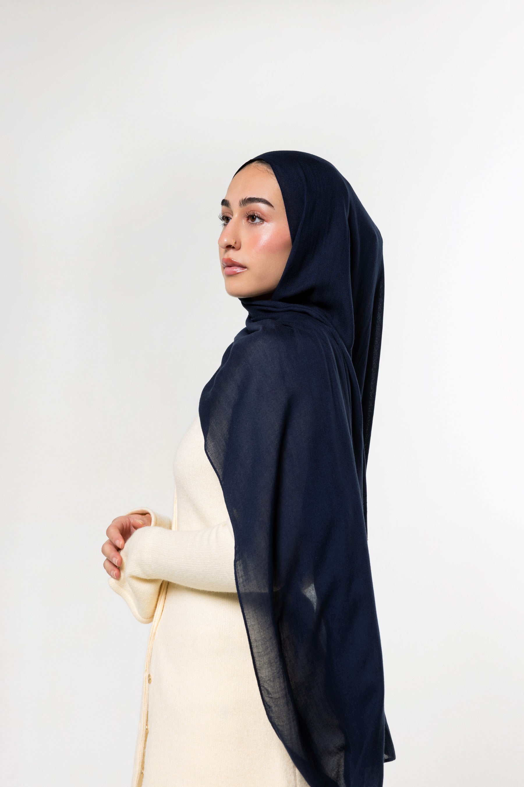 A woman in profile wearing a dark navy blue hijab and a cream-colored dress, photographed against a clean white studio background.