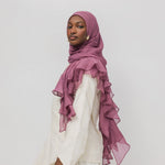 A woman wearing a dusty rose ruffled hijab and a white long-sleeve dress, photographed against a plain light gray studio background.