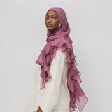 A woman wearing a dusty rose ruffled hijab and a white long-sleeve dress, photographed against a plain light gray studio background.