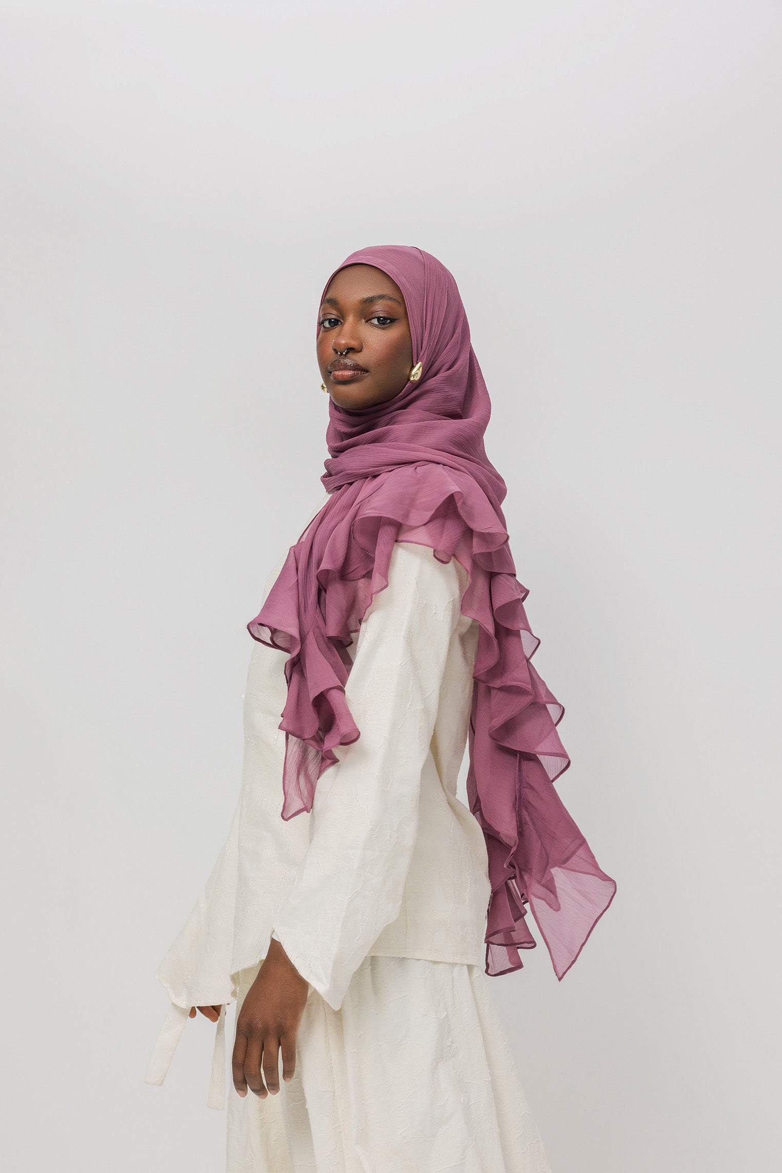 A woman wearing a dusty rose ruffled hijab and a white long-sleeve dress, photographed against a plain light gray studio background.