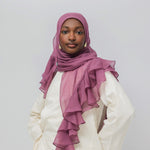 A woman wearing a dusty rose chiffon hijab with layered ruffles and a white tunic, photographed against a neutral light gray background.