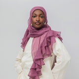 A woman wearing a dusty rose chiffon hijab with layered ruffles and a white tunic, photographed against a neutral light gray background.