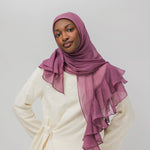 A woman wearing a dusty purple chiffon hijab with layered ruffles and a cream-colored tunic, photographed against a solid light gray background.