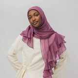 A woman wearing a dusty purple chiffon hijab with layered ruffles and a cream-colored tunic, photographed against a solid light gray background.