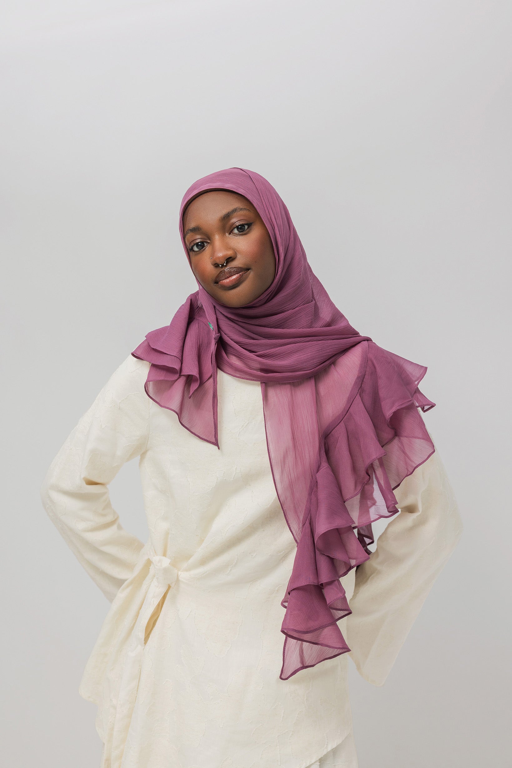 A woman wearing a dusty purple chiffon hijab with layered ruffles and a cream-colored tunic, photographed against a solid light gray background.