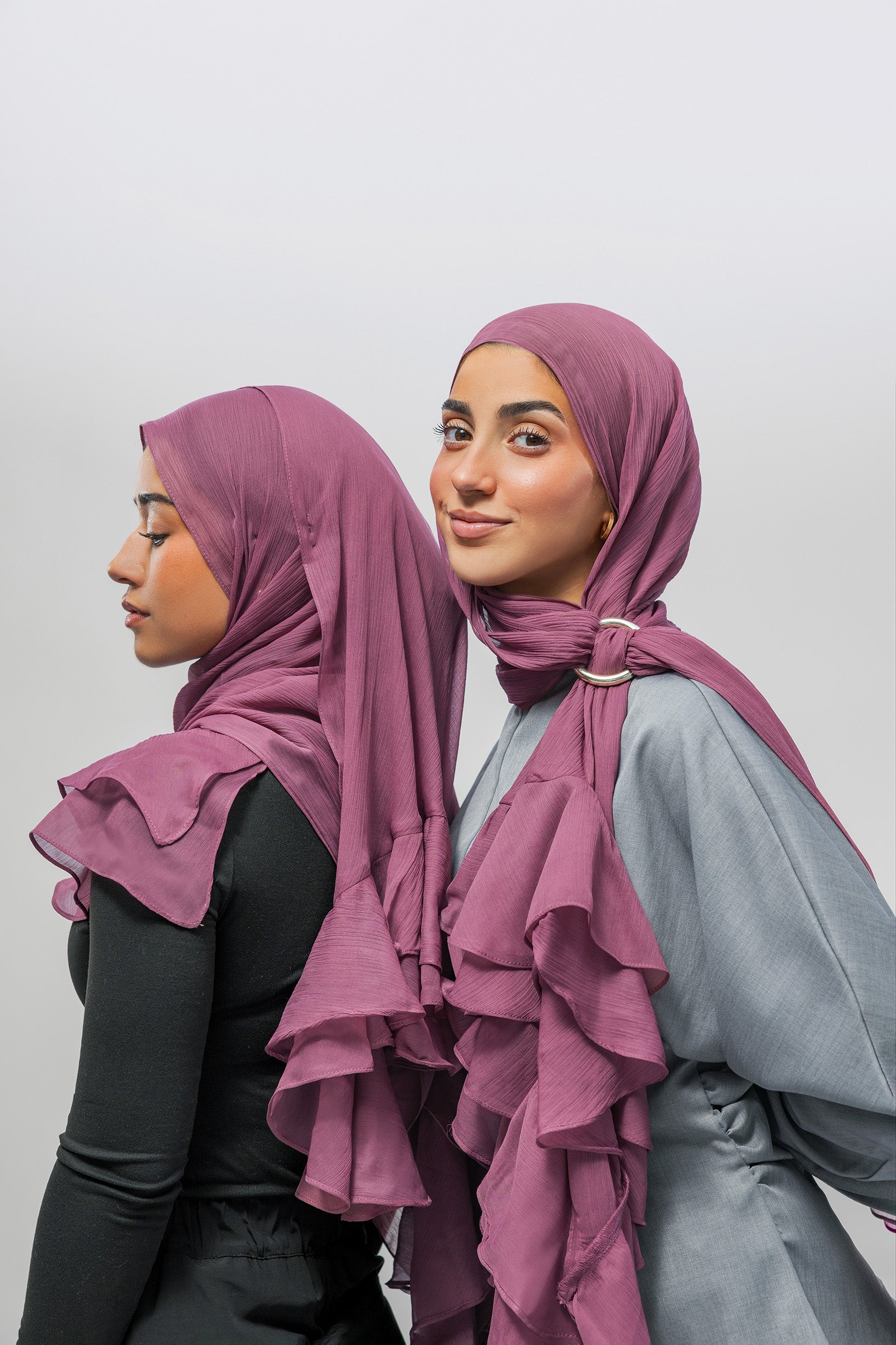 Two women modeling ruffled hijabs in a dusty mauve shade, shown from the side and back against a plain light gray studio background.