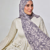 A woman wearing a light purple and white patterned hijab and a cream ribbed long-sleeve top, seated against a plain light gray studio background.