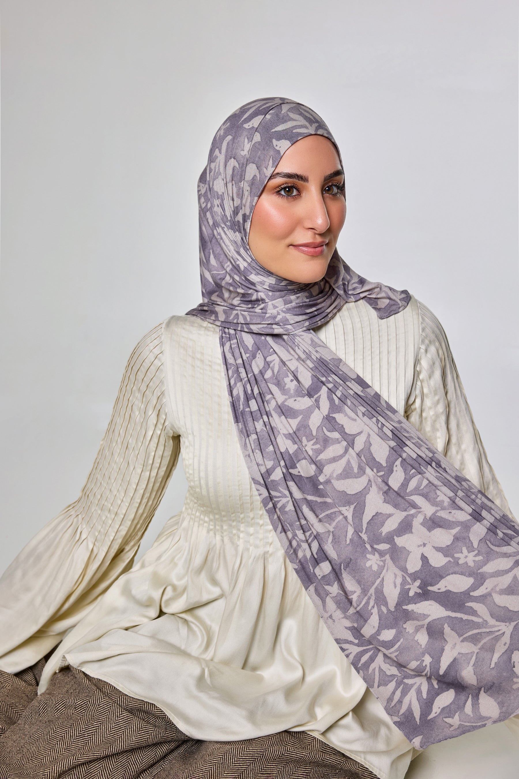 A woman wearing a light purple and white patterned hijab and a cream ribbed long-sleeve top, seated against a plain light gray studio background.
