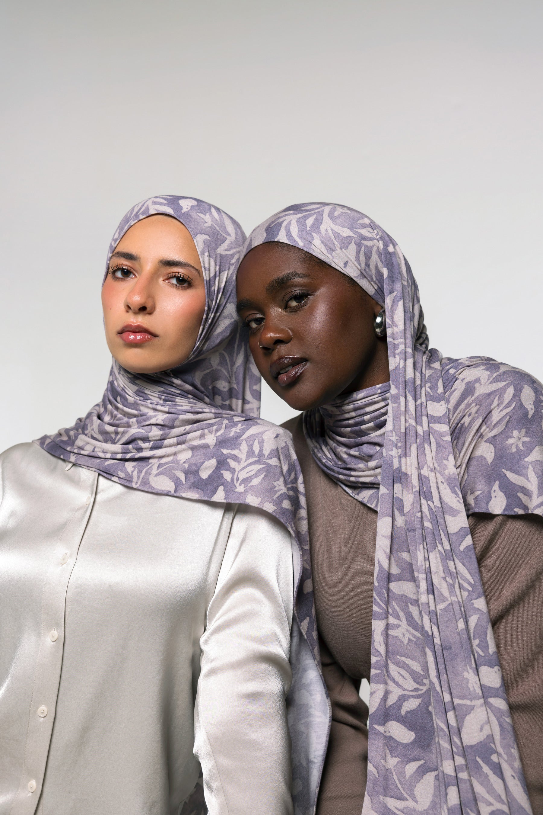 Two women wearing matching patterned hijabs in a studio setting against a light gray background.