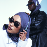 Two models wearing dark sunglasses and matching purple hijabs, one in the foreground adjusting her headphones and the other standing in the background wearing a black leather jacket.
