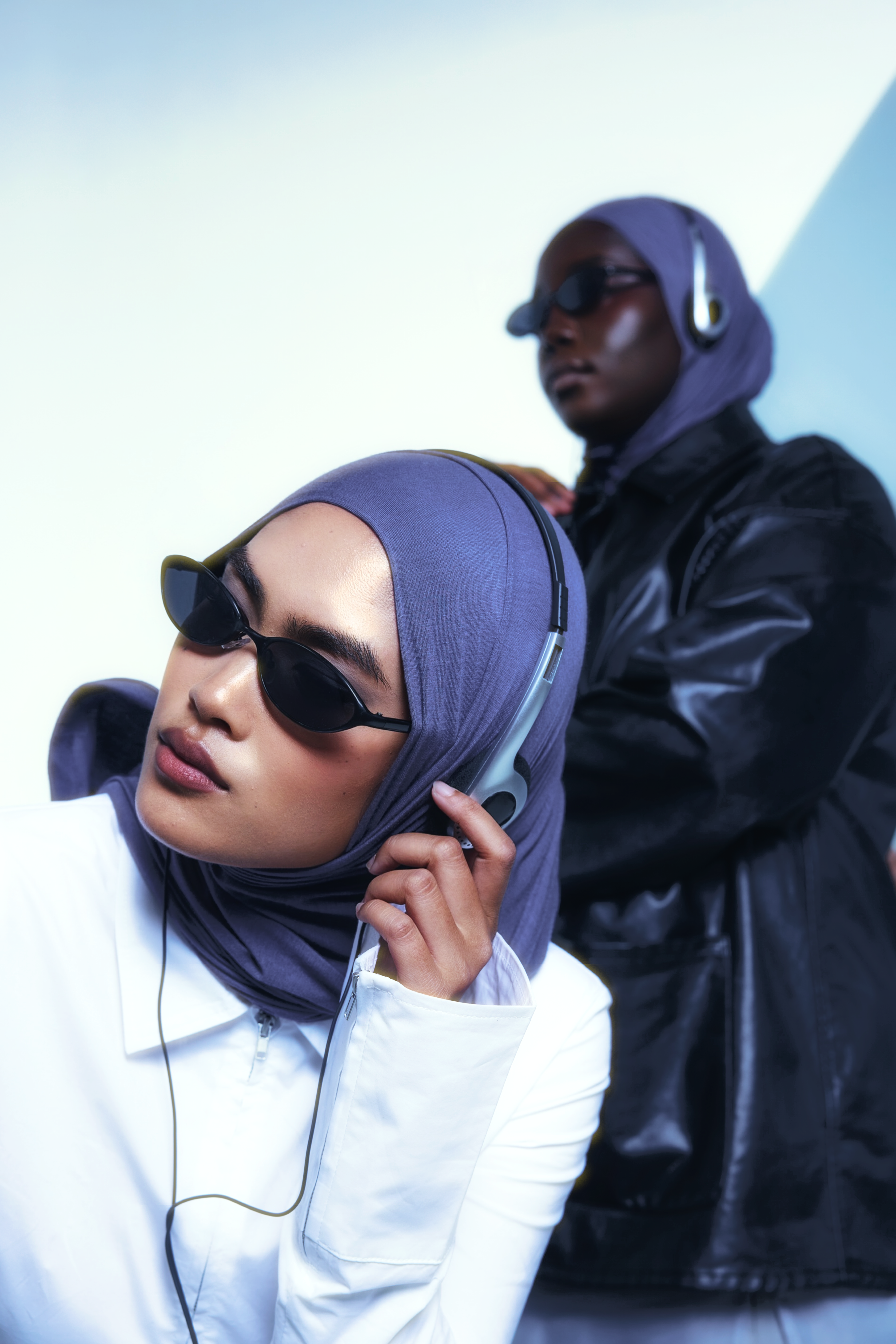 Two models wearing dark sunglasses and matching purple hijabs, one in the foreground adjusting her headphones and the other standing in the background wearing a black leather jacket.