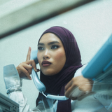 A woman wearing a deep plum purple hijab and a white blouse is shown in a stylized, grainy photograph, holding a light blue landline telephone receiver to her ear.