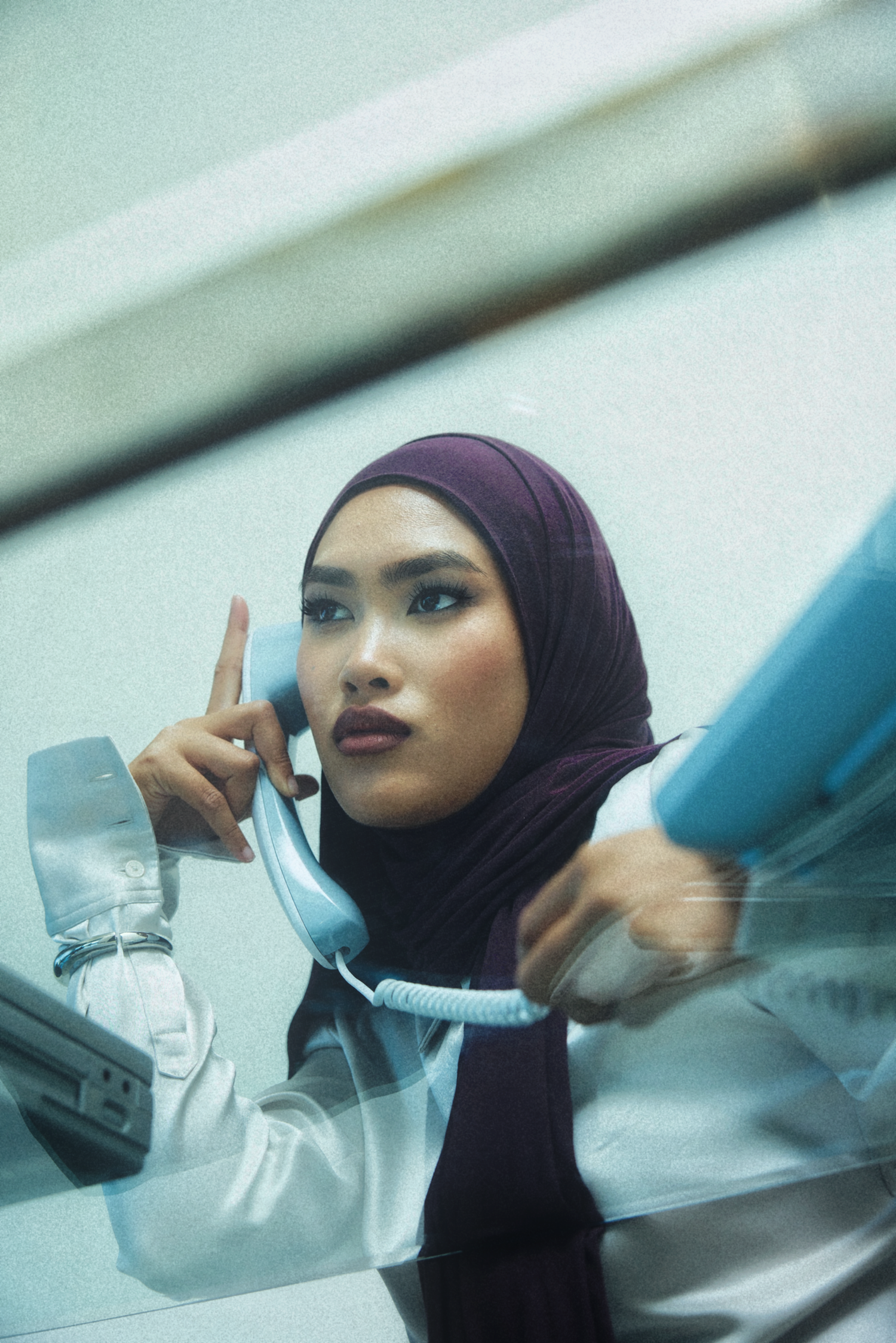 A woman wearing a deep plum purple hijab and a white blouse is shown in a stylized, grainy photograph, holding a light blue landline telephone receiver to her ear.