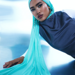 A woman wearing a flowing turquoise hijab and a dark navy top, posed against a cool-toned blue and white studio background.