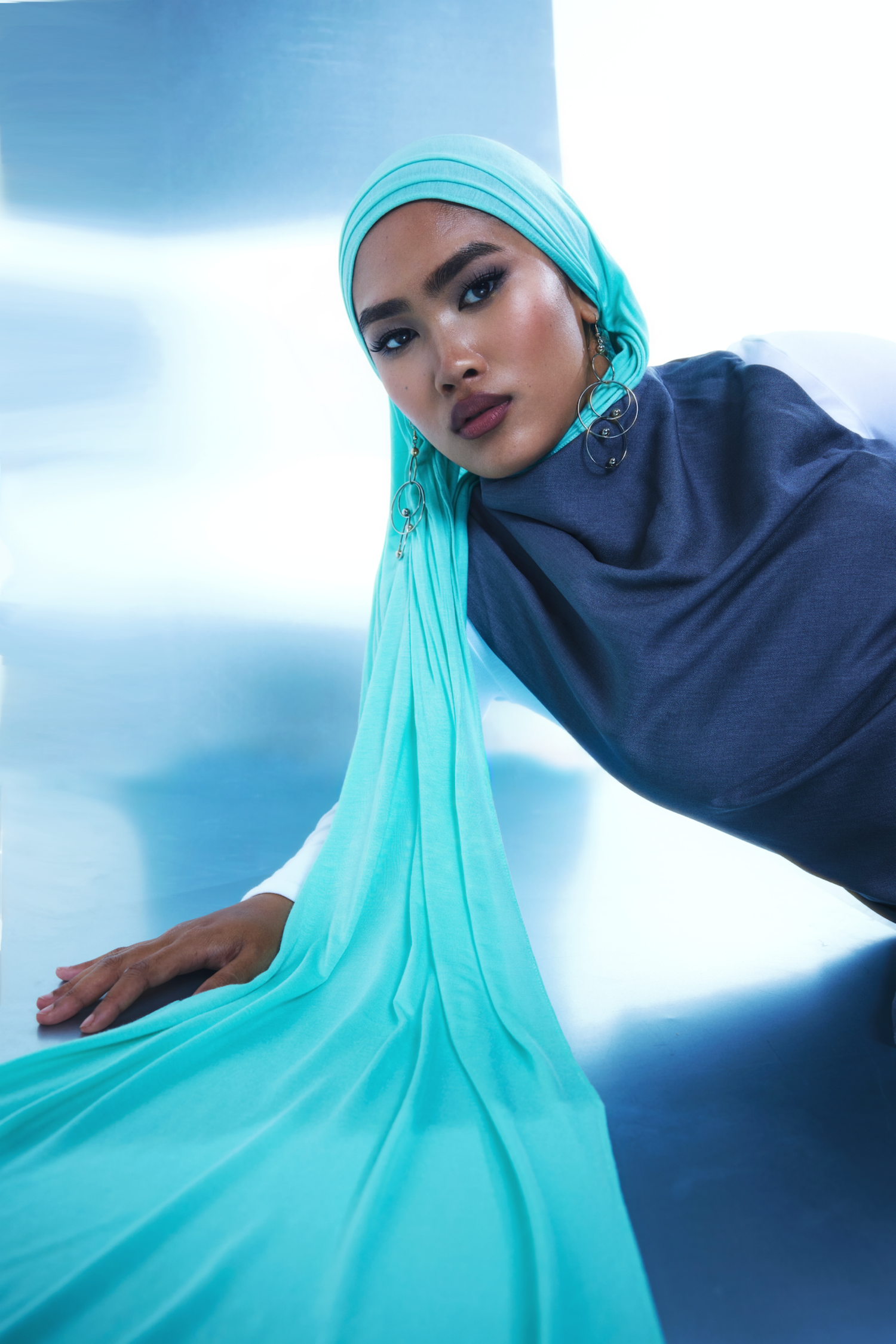 A woman wearing a flowing turquoise hijab and a dark navy top, posed against a cool-toned blue and white studio background.