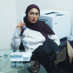 A woman wearing a maroon hijab and glasses sits in an office environment, holding a white landline telephone receiver. She is dressed in a white long-sleeve blouse, dark trousers, and black high-heeled boots, with a glass desk in the foreground and a printer in the background.