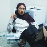 A woman wearing a maroon hijab and glasses sits in an office environment, holding a white landline telephone receiver. She is dressed in a white long-sleeve blouse, dark trousers, and black high-heeled boots, with a glass desk in the foreground and a printer in the background.