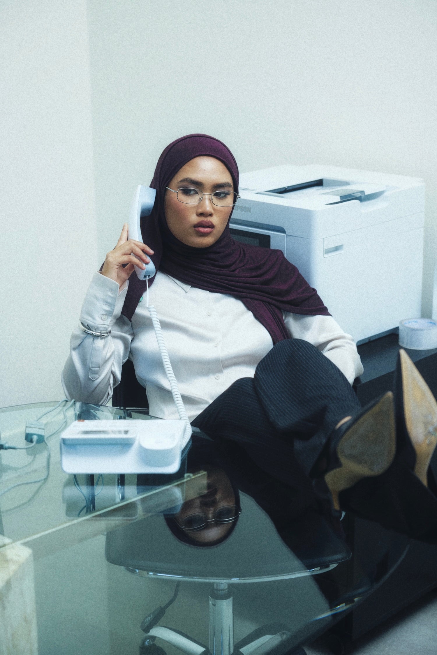 A woman wearing a maroon hijab and glasses sits in an office environment, holding a white landline telephone receiver. She is dressed in a white long-sleeve blouse, dark trousers, and black high-heeled boots, with a glass desk in the foreground and a printer in the background.