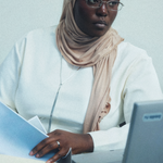A woman wearing a beige hijab and glasses, dressed in a white top, sitting at a desk with a laptop and holding papers.